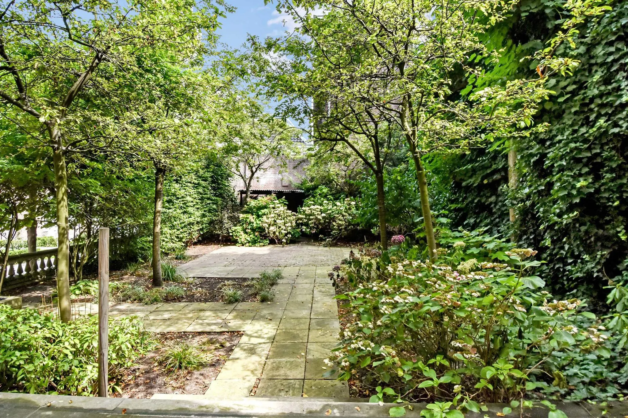 Groene, beschutte stadstuin aan het Lange Voorhout met betegeld pad, bloeiende struiken en schaduwrijke bomen.